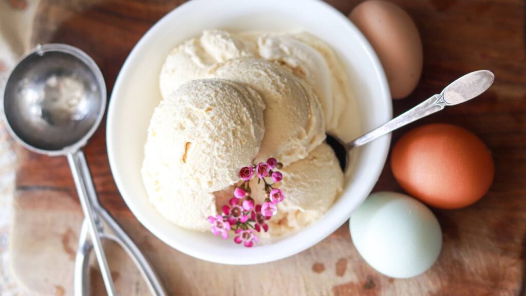 A bowl of homemade vanilla ice cream with pink flowers, eggs, and an ice cream scoop.