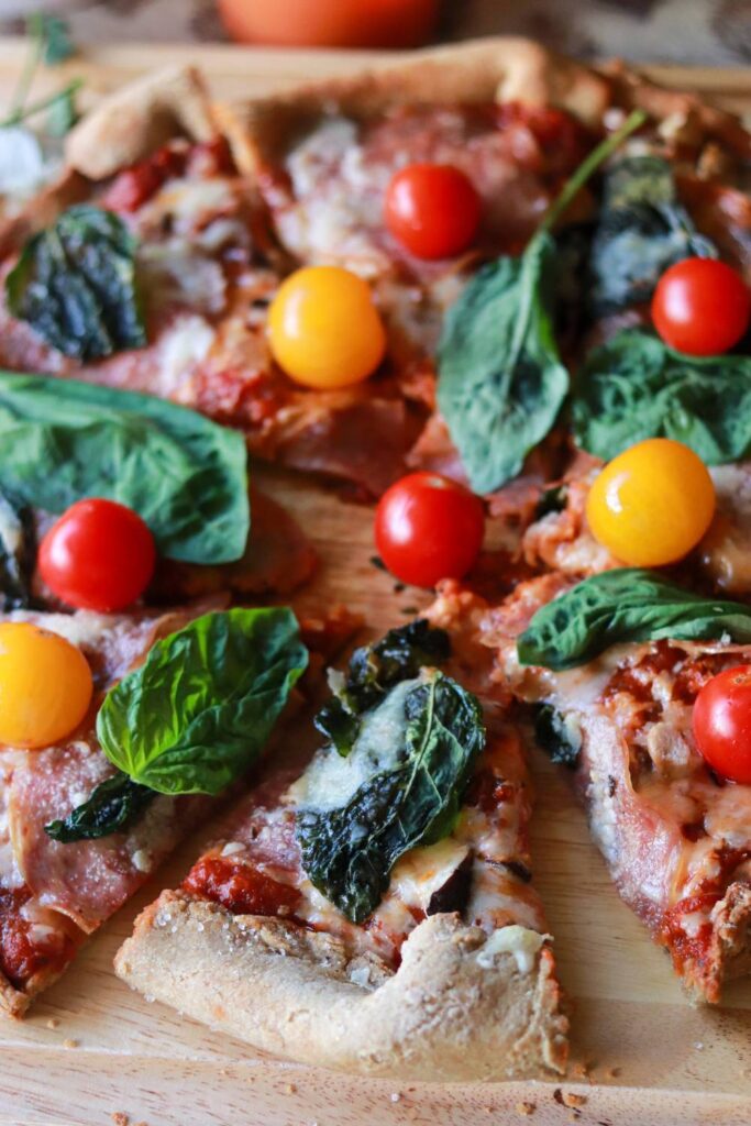 Close-up of a sliced homemade freshly milled sourdough pizza topped with cherry tomatoes, fresh basil leaves, and melted cheese on a wooden cutting board.