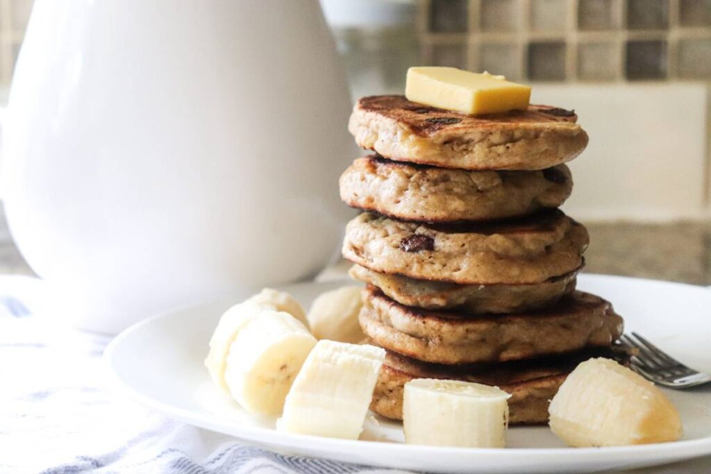 Stack of fluffy sourdough discard banana pancakes with butter on top, served with sliced bananas on a white plate