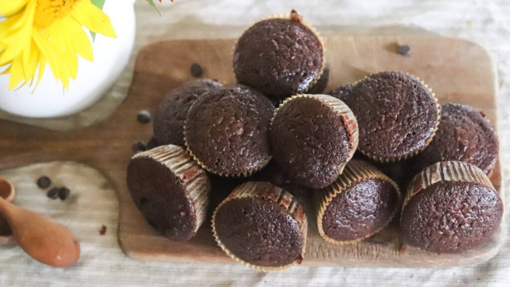 Pile of chocolate sourdough discard muffins on a wooden board next to a white vase.