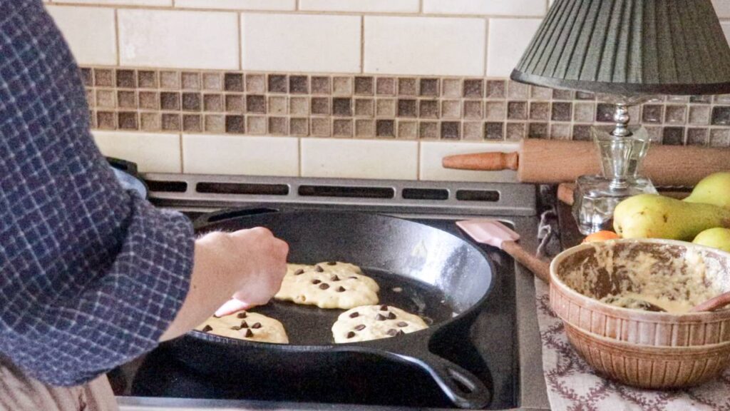 a woman standing at a stovetop cooking sourdough pancakes on a hot cast iron skillet.