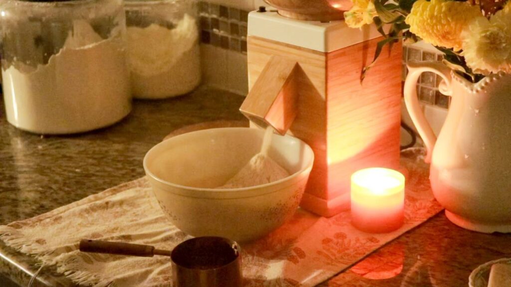 a kitchen lighted by candlelight with a grain mill sitting on the counter milling fresh flour into a large mixing bowl.