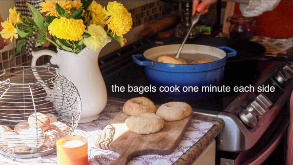 three sourdough bagels sitting out on a wooden cutting board on a kitchen counter with bagels being cooked on the stovetop nearby.