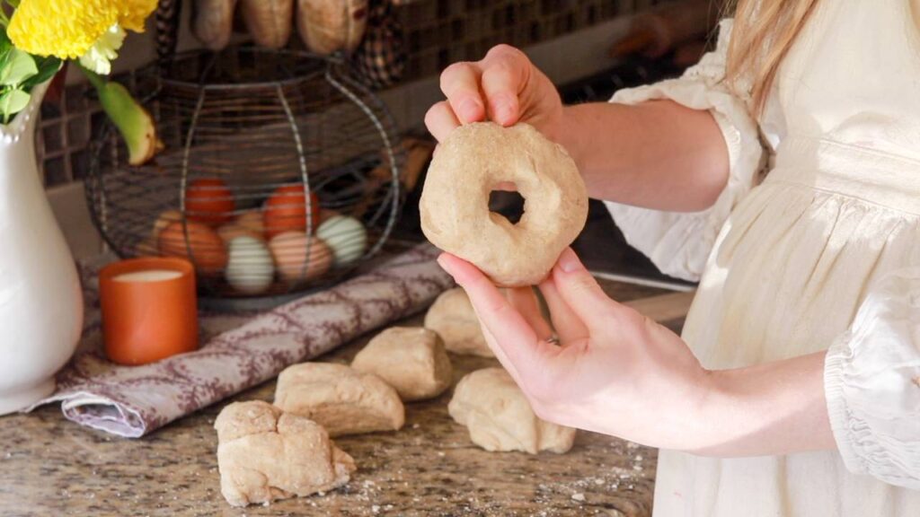 woman holding up a sourdough bagel dough that has been rolled and made into a donut shape with other dough ready to be worked in the background.