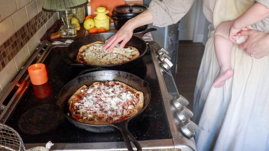 Woman making sourdough pizza while holding a baby on her hip, spreading cheese and toppings onto pizzas in cast iron pans on a stove.