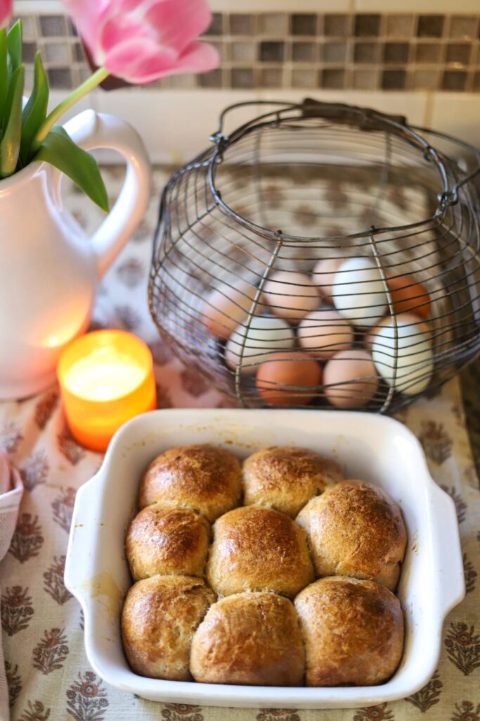 Baked sourdough rolls in a dish next to fresh eggs, flowers, and a candle