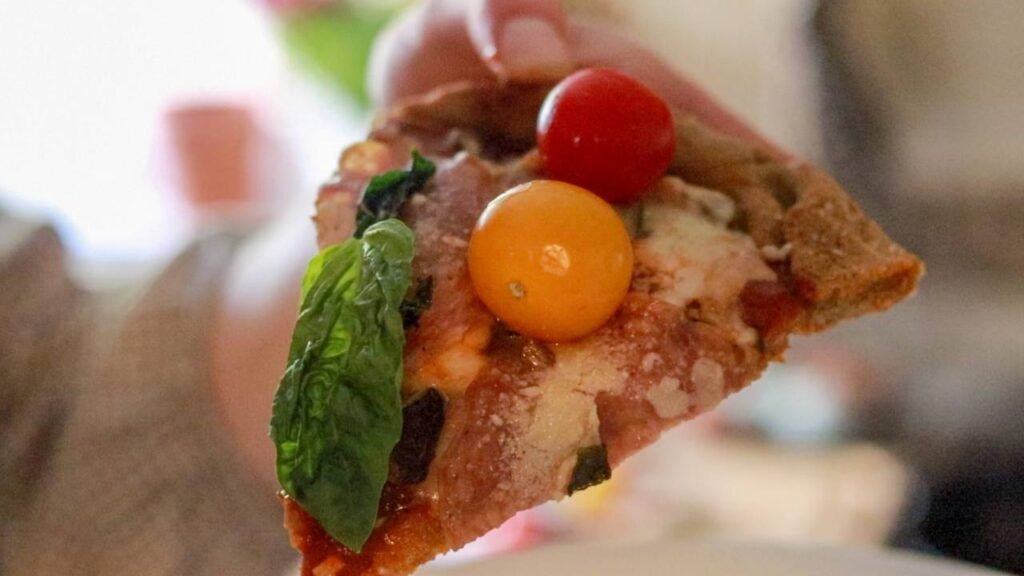 Close-up of a single slice of homemade sourdough pizza held up, topped with red and yellow cherry tomatoes, melted cheese, and fresh basil.