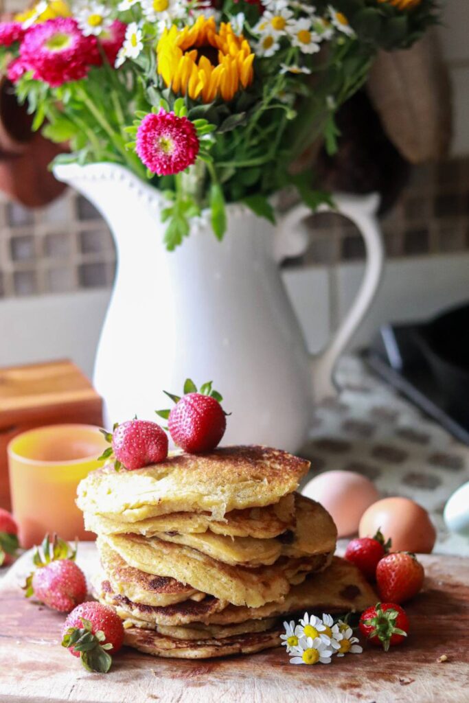 Buttermilk sourdough pancakes stacked on top of each other on a cutting board with whole strawberries on top and around the stack as well as little white daisies and a tall white vase with bright flowers.