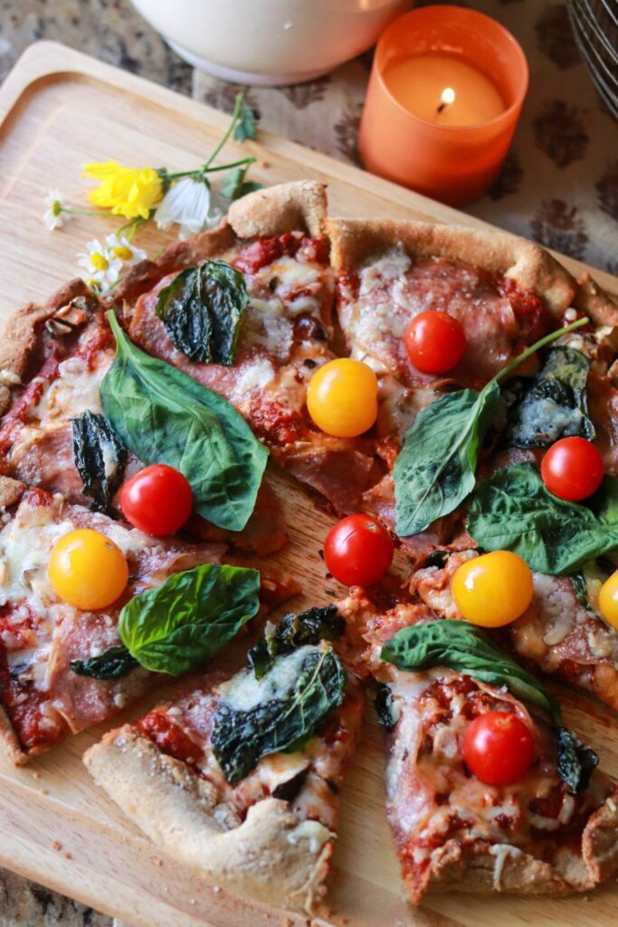 Overhead view of a homemade pizza with colorful cherry tomatoes and basil, surrounded by daisies, a lit candle, and cozy kitchen decor.