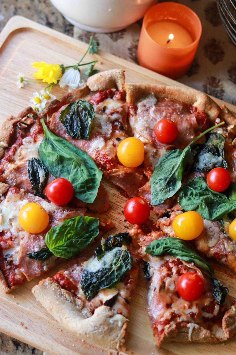 Overhead view of a homemade pizza with colorful cherry tomatoes and basil, surrounded by daisies, a lit candle, and cozy kitchen decor.