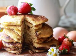 a tall stack of buttermilk pancakes on a cutting board with white daisies and strawberries placed around it and on top of the pancakes, with a sliver taken out of the stack.
