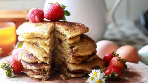 a tall stack of buttermilk pancakes on a cutting board with white daisies and strawberries placed around it and on top of the pancakes, with a sliver taken out of the stack.