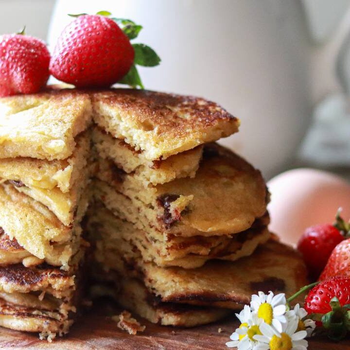 a tall stack of buttermilk pancakes on a cutting board with white daisies and strawberries placed around it and on top of the pancakes, with a sliver taken out of the stack.