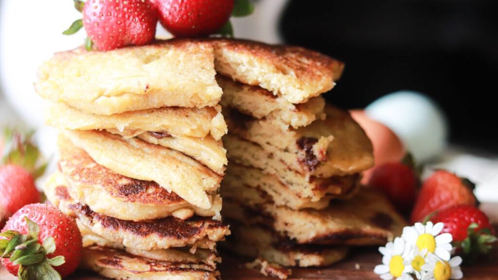 a tall stack of buttermilk pancakes on a cutting board with strawberries placed around it and on top of the pancakes, with a sliver taken out of the stack.