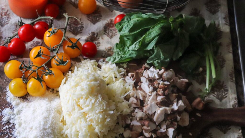 Assortment of fresh pizza ingredients including shredded cheese, chopped mushrooms, yellow and red cherry tomatoes, and fresh basil.