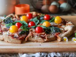 Front view of a whole pizza with one slice missing, sitting on a wooden board with daisies and an orange candle in the background.