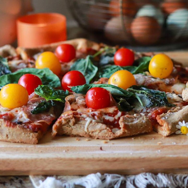 Front view of a whole pizza with one slice missing, sitting on a wooden board with daisies and an orange candle in the background.
