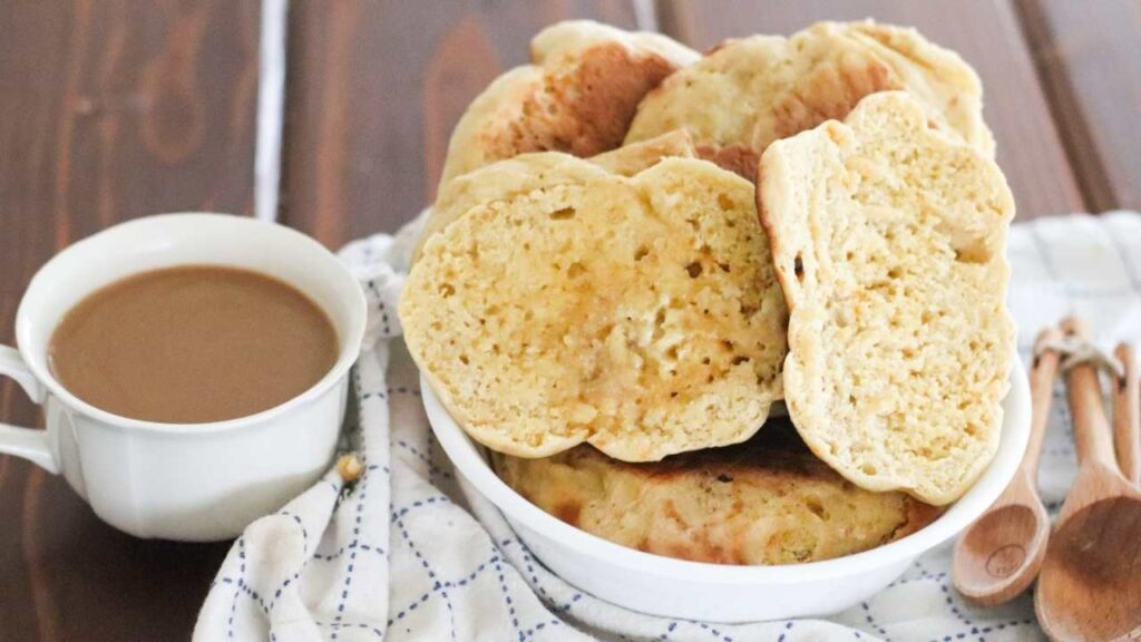 Homemade einkorn sourdough English muffins stacked in a bowl, served with a cup of coffee on the side.