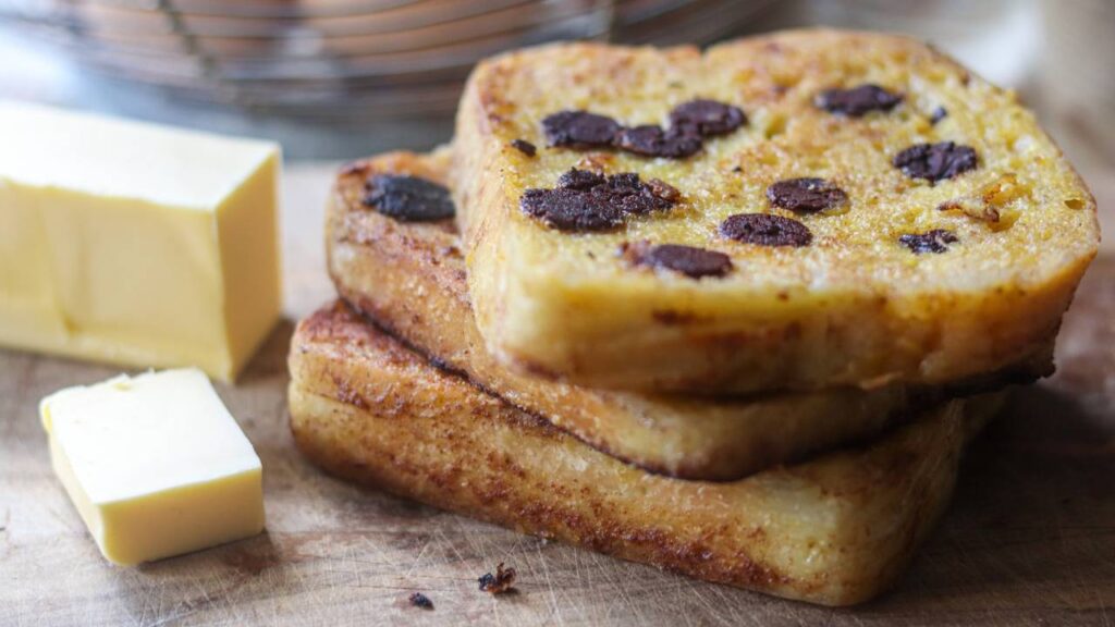 Thick slices of chocolate chip sourdough French toast stacked on a wooden board with butter on the side.
