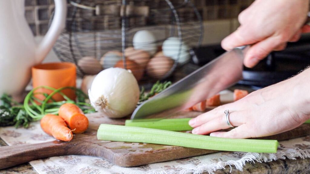 Person slicing celery on a wooden board surrounded by chowder ingredients.
