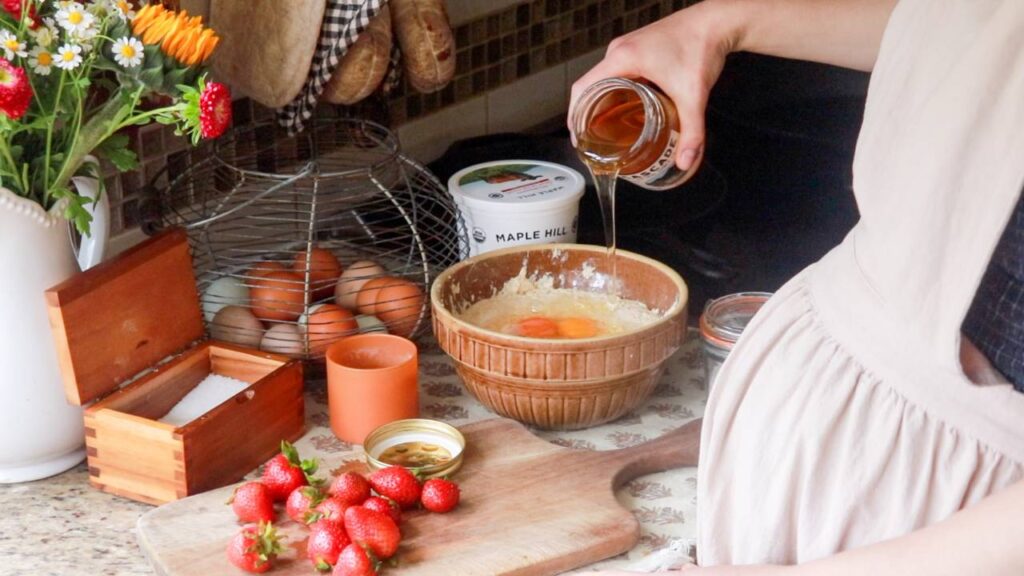 a woman wearing a white apron standing at a kitchen counter pouring honey from a jar into a mixing bowl with pancake ingredients.