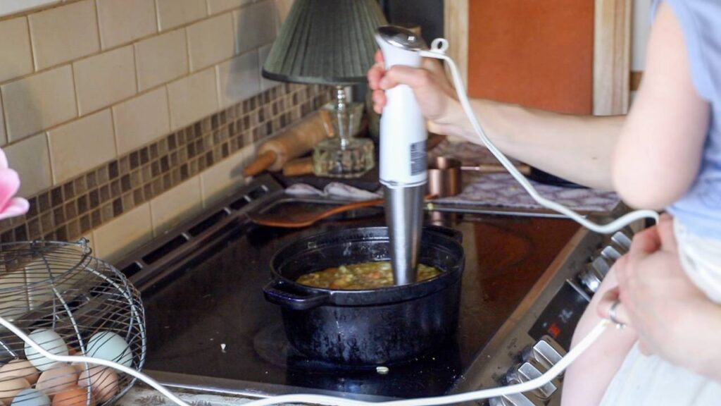 Using an immersion blender to blend soup in a black Dutch oven on the stove while holding a child.
