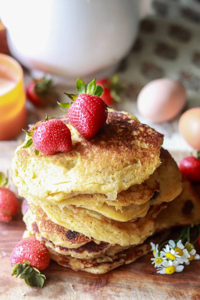 Freshly milled sourdough pancakes stacked on top of each other on a cutting board with whole strawberries on top and around the stack as well as little white daisies.