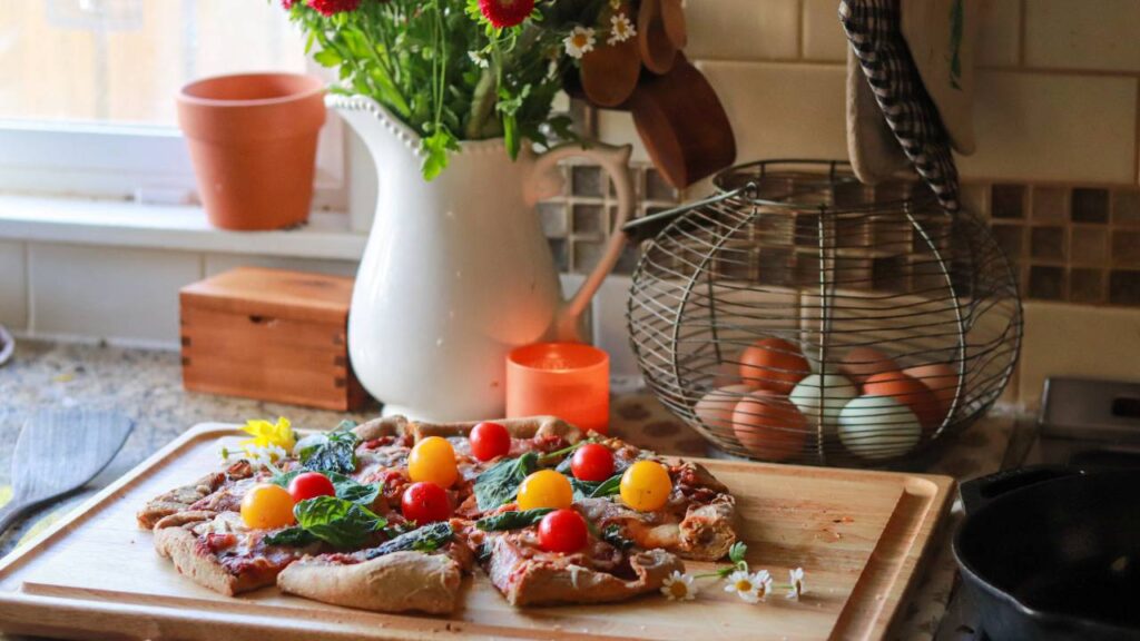 Cozy kitchen with a pizza on a cutting board, daisies, a white pitcher of flowers, a candle, and a wire basket of eggs in the background.