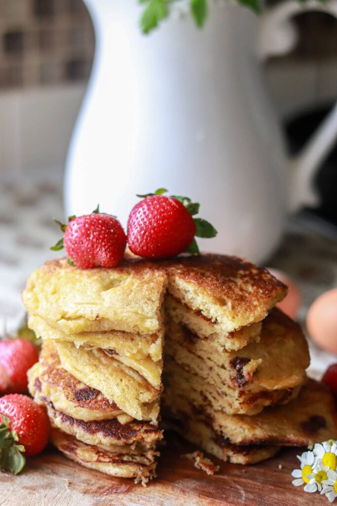 a tall stack of buttermilk pancakes on a cutting board with strawberries placed around it and on top of the pancakes, with a sliver taken out of the stack.