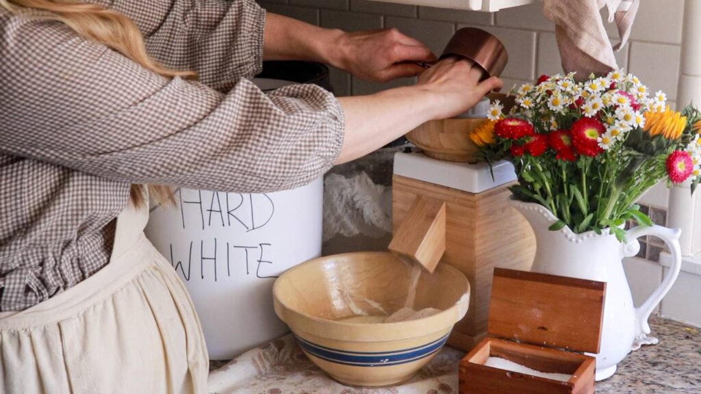 Woman in vintage-style apron scooping freshly ground flour from a container beside a mixing bowl and fresh flowers.