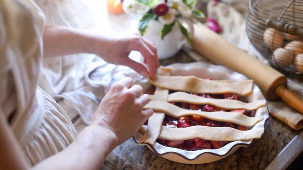 Hands placing lattice strips of pie crust over a strawberry pie.