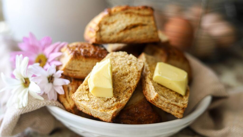 Flaky homemade biscuits with butter in a white bowl and fresh flowers.