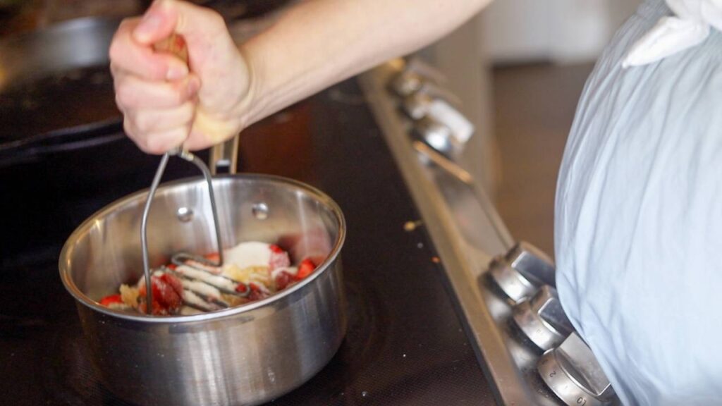 Hand mashing strawberries and sugar in a saucepan on the stove.