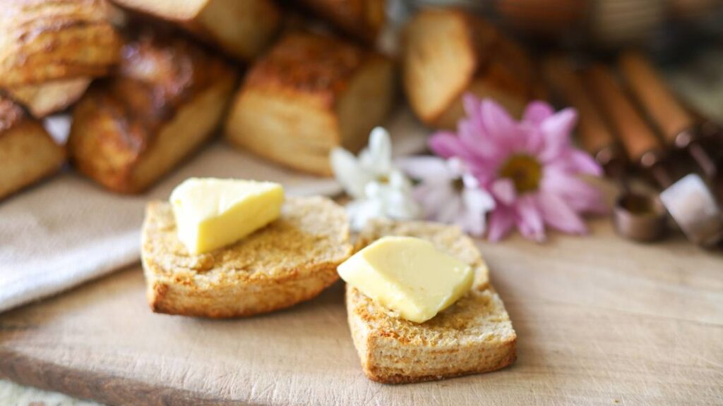 Two buttered biscuits on a wooden board with flowers in the background.