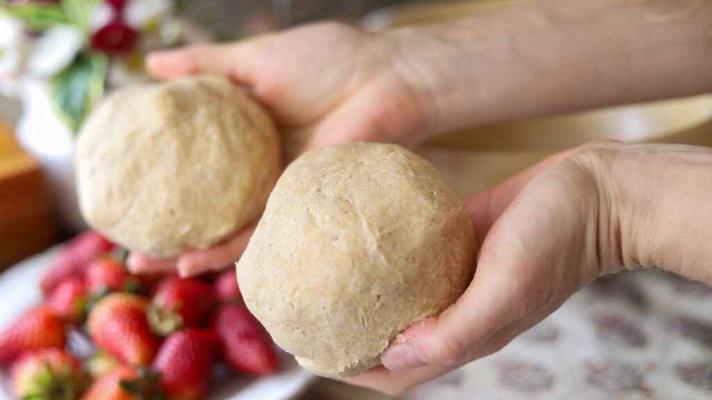 Two hands holding dough balls with strawberries in the background.
