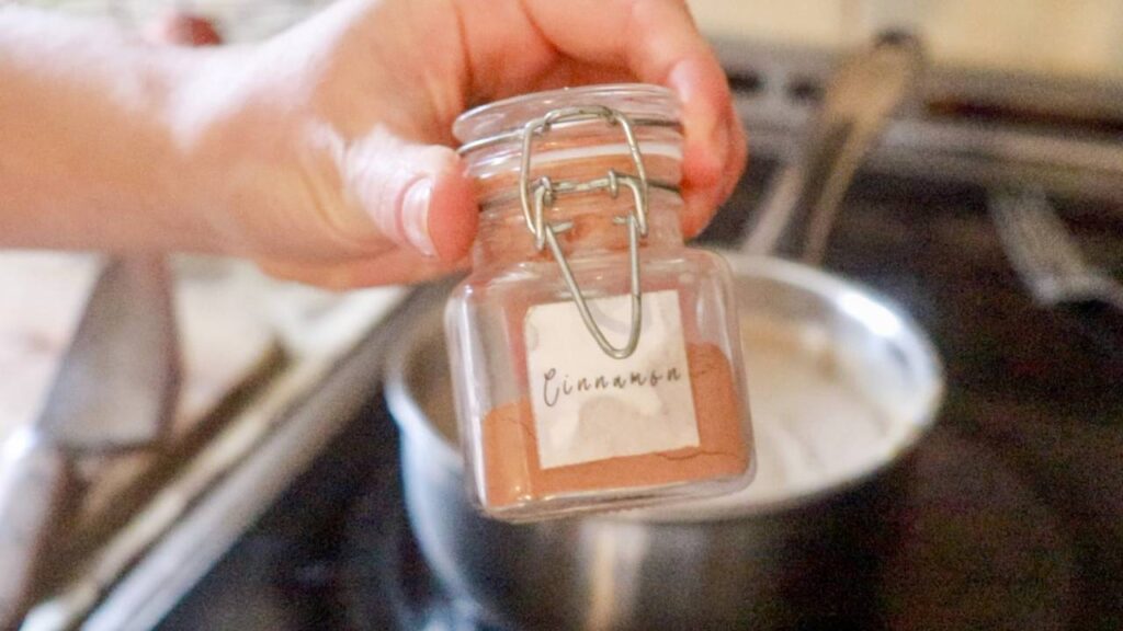 Hand holding a jar of cinnamon over a pot on the stove.