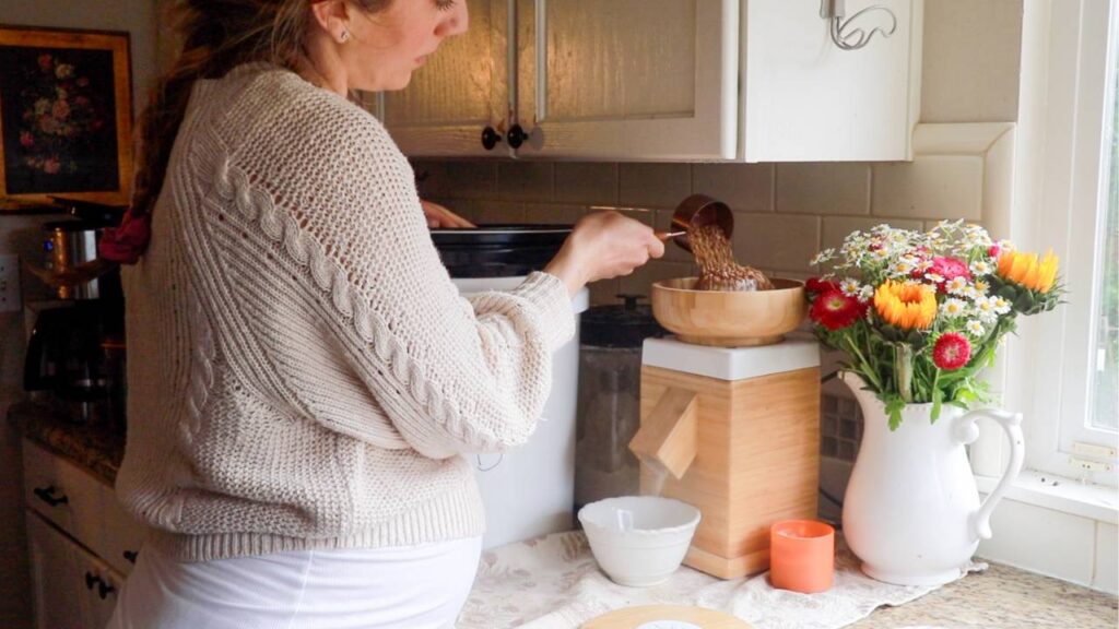 Woman pouring grains into a grain mill in a cozy kitchen.