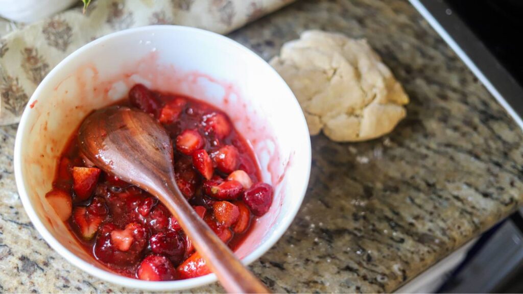 Strawberry filling in a mixing bowl with a wooden spoon and pie dough on the counter.