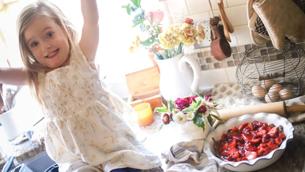 Little girl in a cream dress smiling next to an unbaked strawberry pie on the counter.