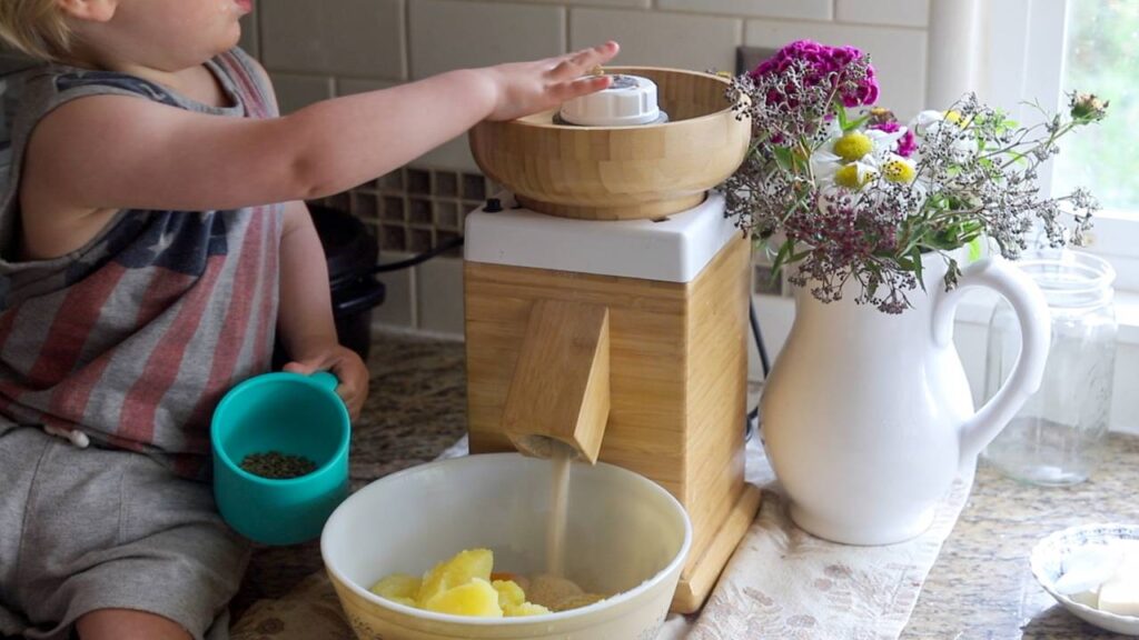 Child helping pour grain into a flour mill next to a bowl of potatoes.