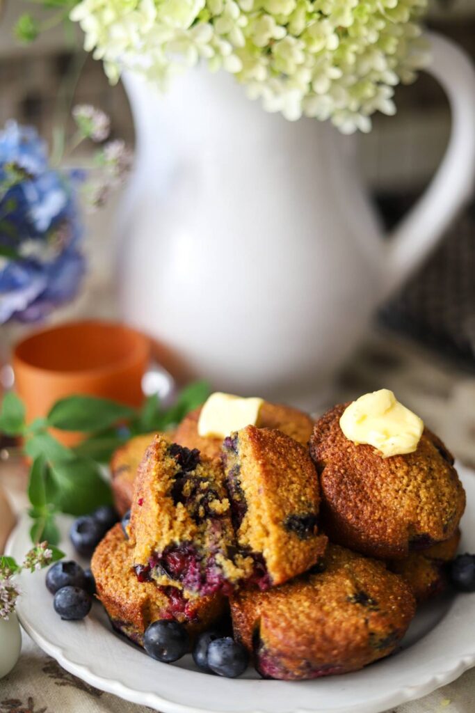 Blueberry muffins on a plate with butter on top and flowers in the background.