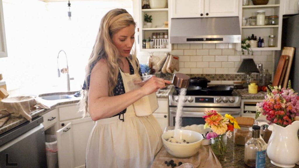 Woman pouring sugar into a bowl of blueberries in a cozy kitchen