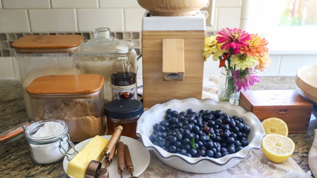 Ingredients for blueberry cobbler laid out on the counter, including blueberries, flour, butter, and a grain mill