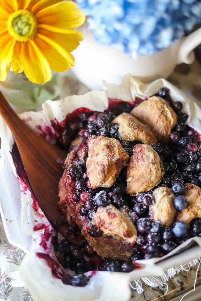 Baked blueberry cobbler in a white dish with visible biscuit topping and a wooden spoon