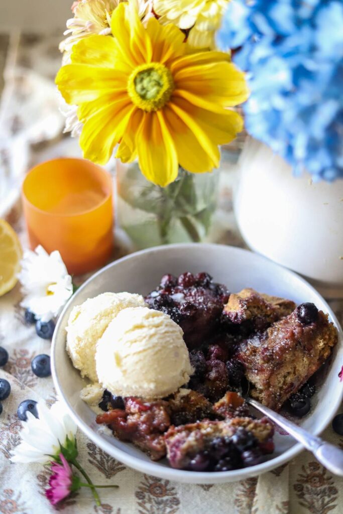 Bowl of blueberry cobbler served with vanilla ice cream and fresh flowers in the background