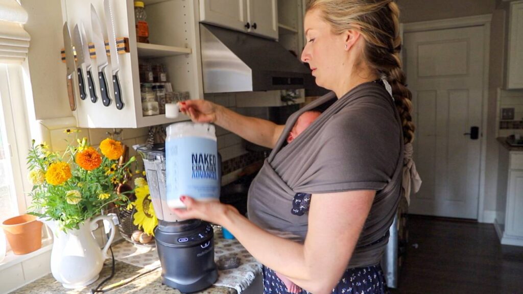 Mom wearing baby in wrap while scooping collagen powder into blender