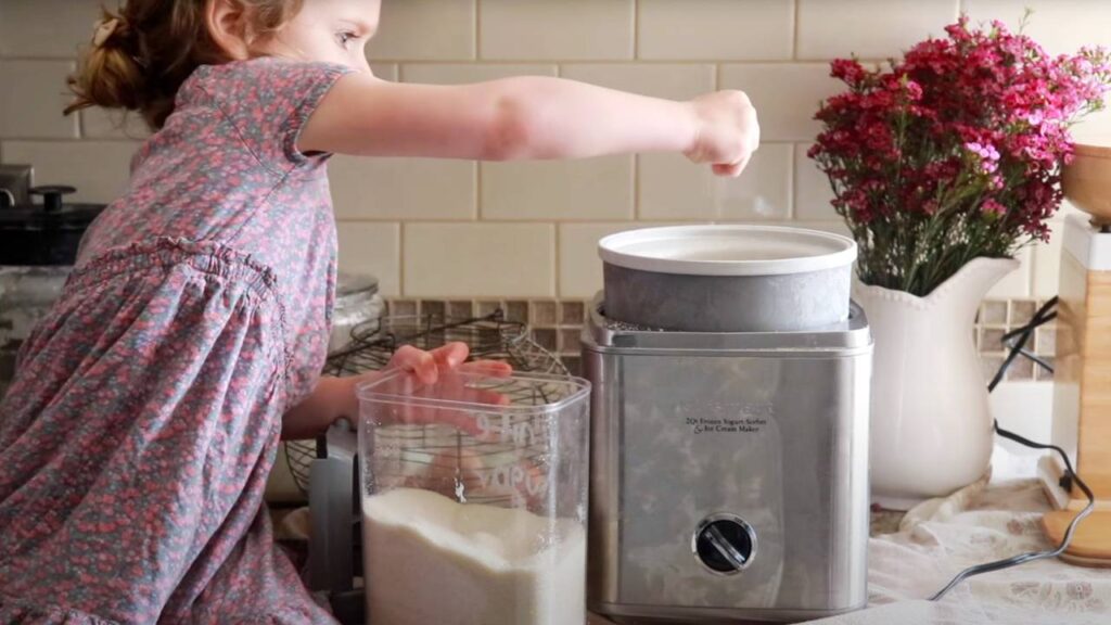 A little girl pouring sugar into an ice cream maker on a kitchen counter.