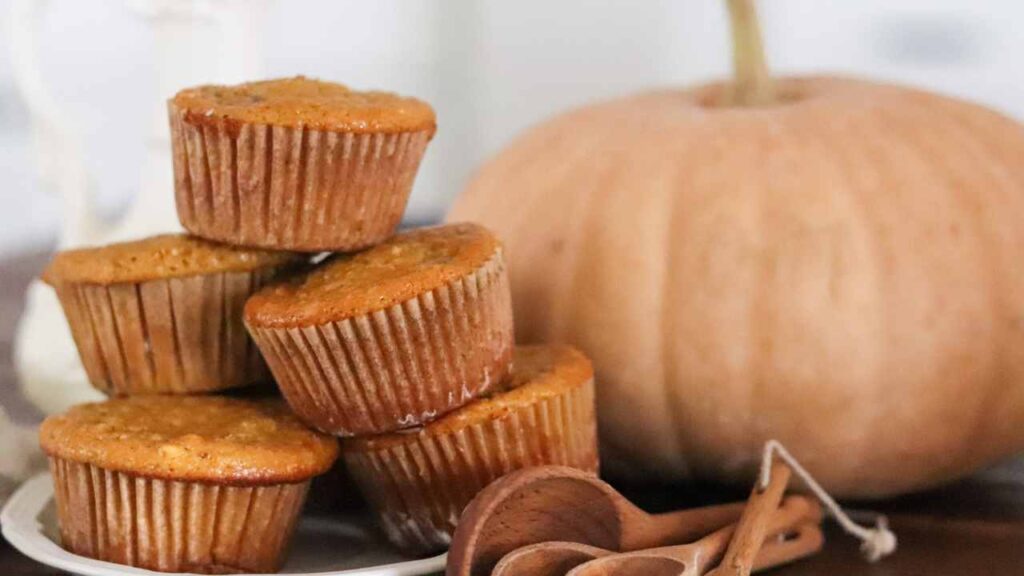 a stack of fresh pumpkin muffins with measuring spoons sitting nearby and a pumpkin in the background.