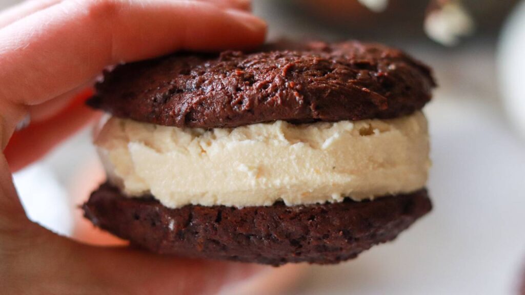 Hand holding a chocolate cookie ice cream sandwich with vanilla ice cream.