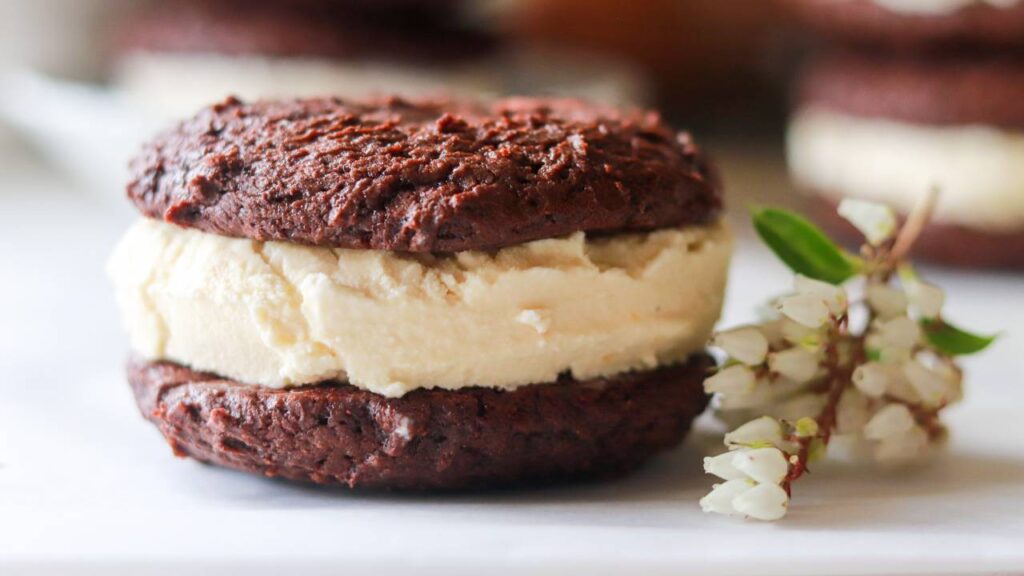 Close-up of a homemade chocolate cookie ice cream sandwich beside white flowers.